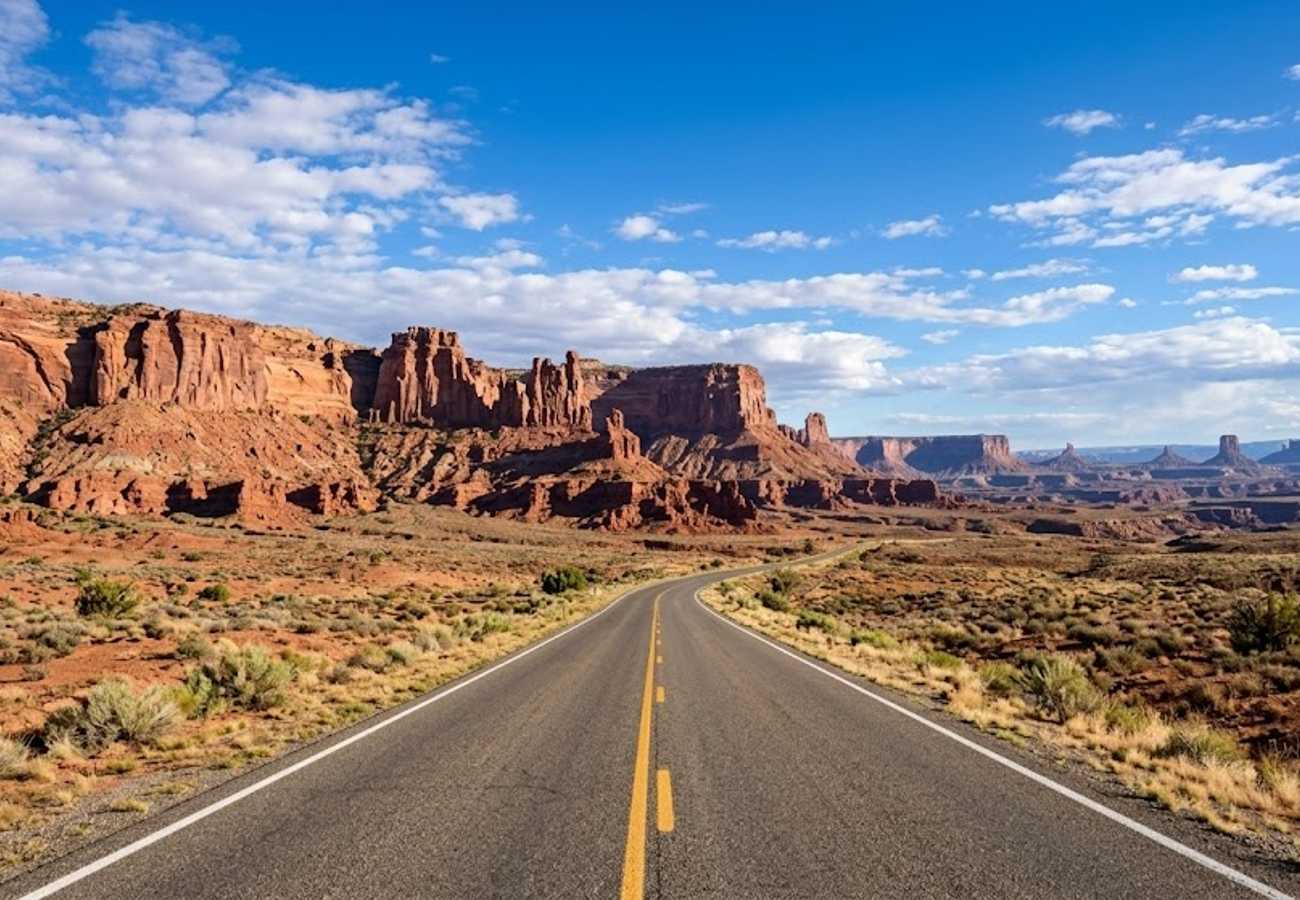 Open paved desert highway stretching toward massive red sandstone cliffs and hoodoos under a brilliant Southwest sky