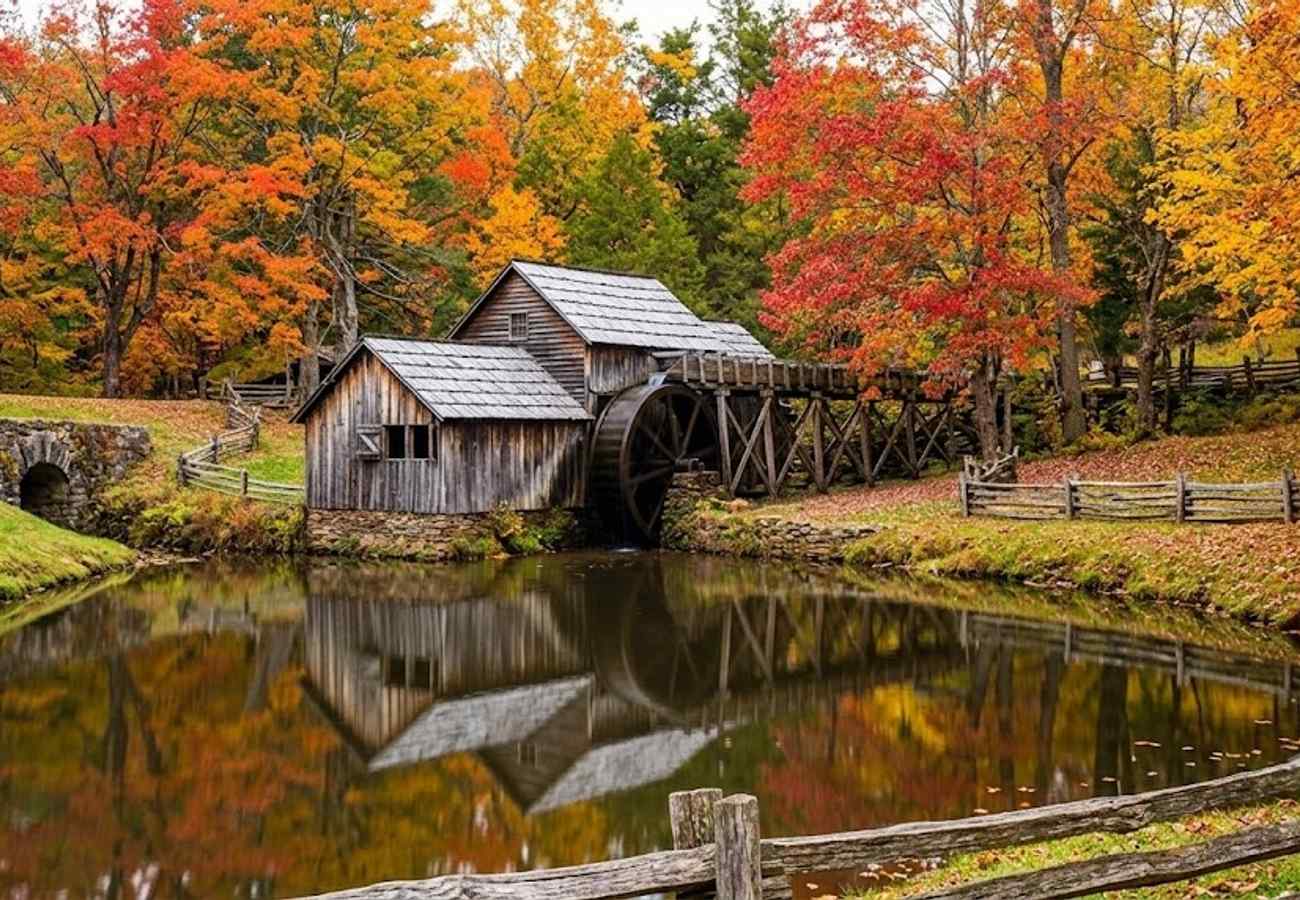 Historic water-powered wooden gristmill with water wheel reflecting in a tranquil pond surrounded by autumn leaves