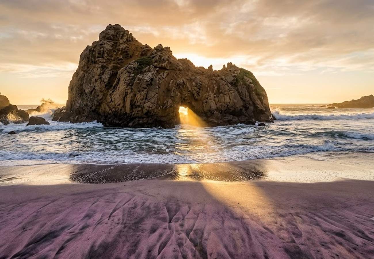 Natural Keyhole rock arch on Pfeiffer Beach with sunlight beaming through the sea portal onto purple garnet sand and crashing waves